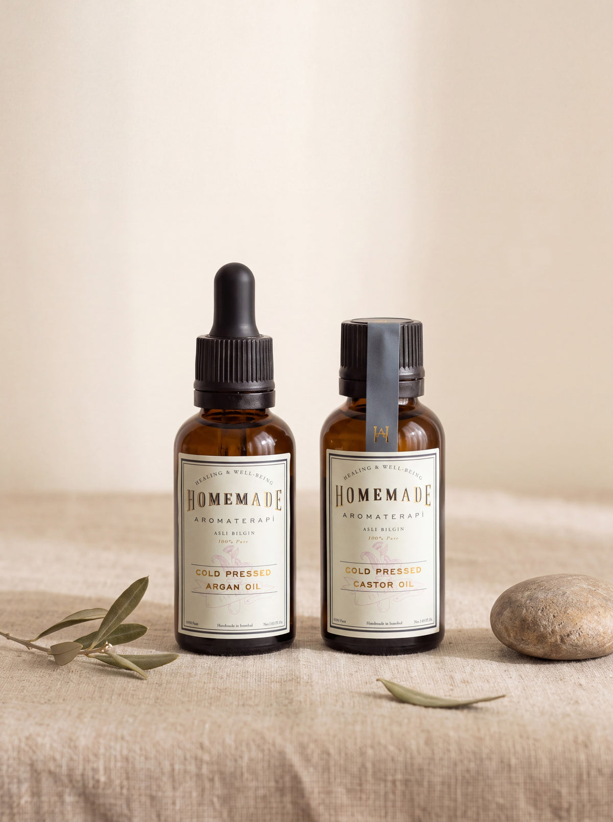 Two HomeMade Aromaterapi cold-pressed oil bottles (Argan and Castor) displayed on a neutral fabric with olive branches and a smooth stone.