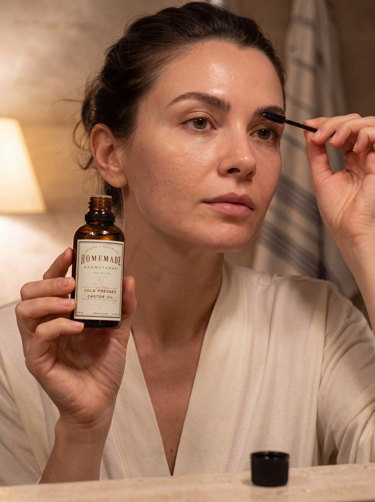 A woman applies makeup while holding a bottle of HMA Extra Virgin Cold Pressed Castor Oil in a warm, indoor setting.