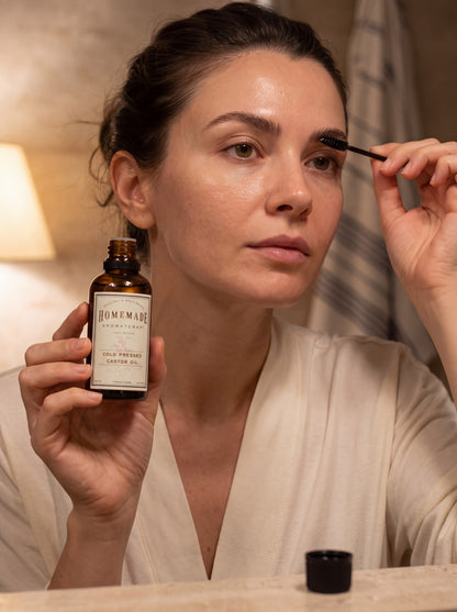 A woman applies makeup while holding a bottle of HMA Extra Virgin Cold Pressed Castor Oil in a warm, indoor setting.