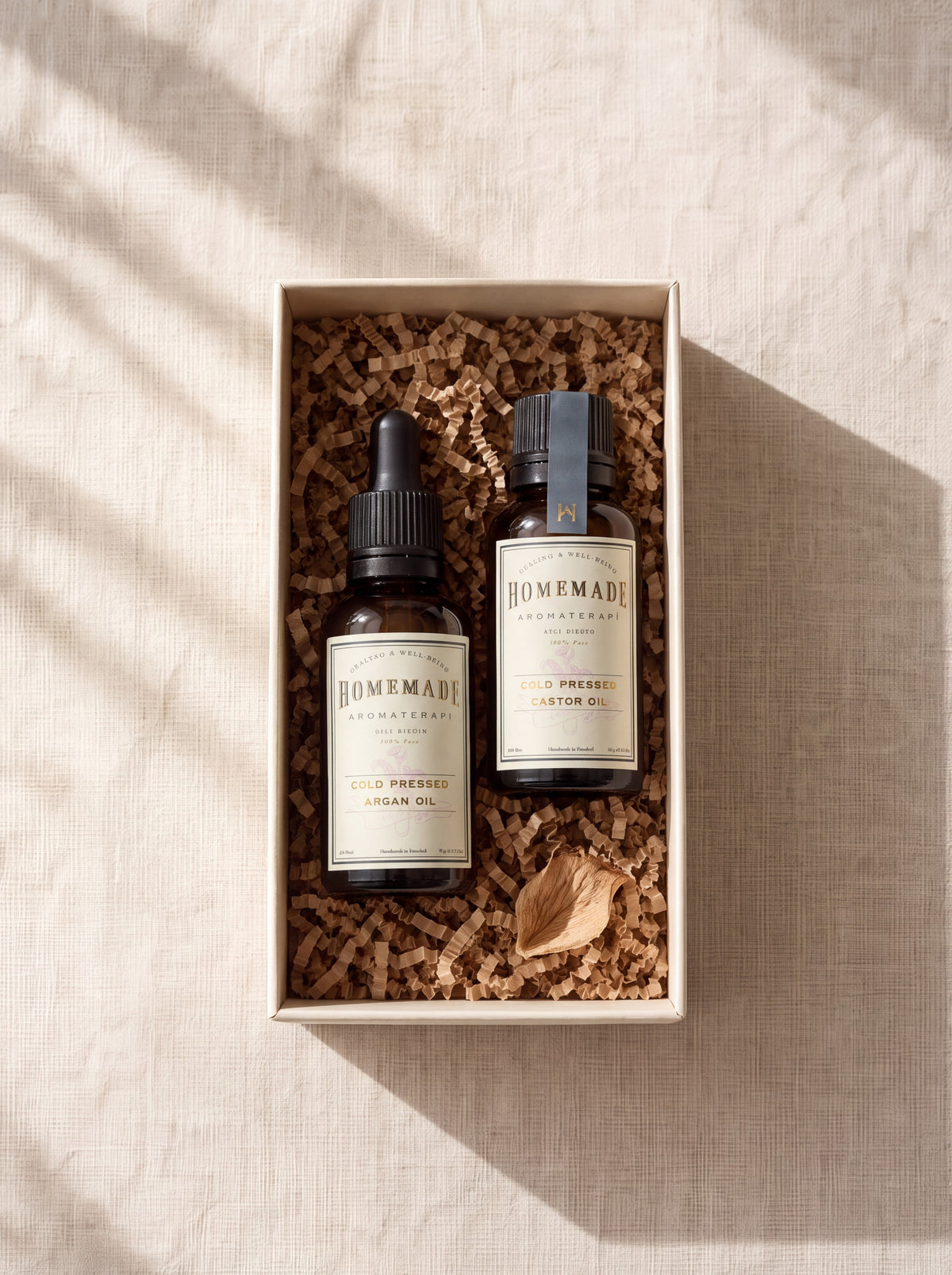 An open gift box with cork fill containing two HomeMade Aromaterapi cold-pressed oil bottles labeled Argan Oil and Castor Oil with a decorative dried flower.