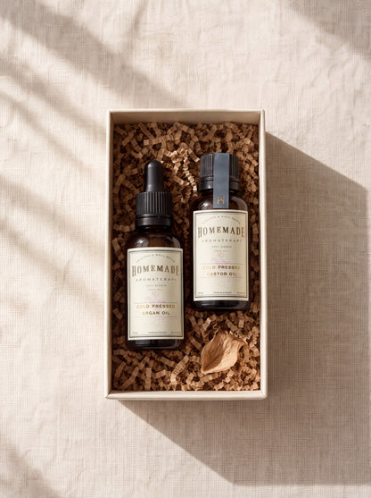 An open gift box with cork fill containing two HomeMade Aromaterapi cold-pressed oil bottles labeled Argan Oil and Castor Oil with a decorative dried flower.